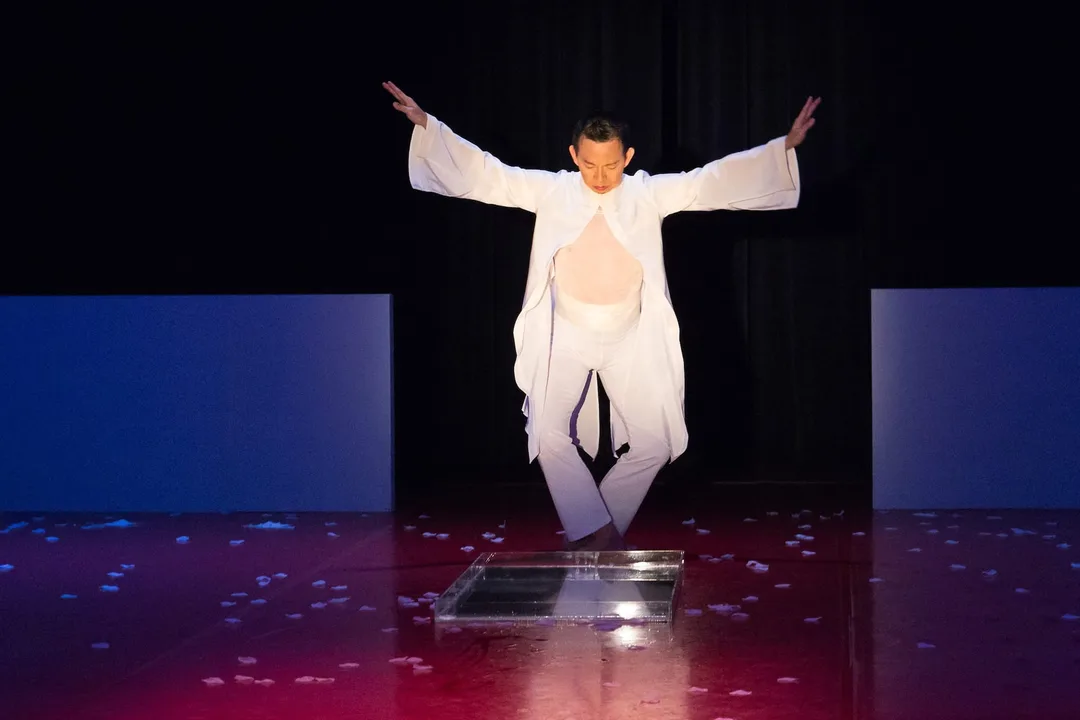 Minh is on stage in all white, gazing at something on the floor. His legs form a diamond shape and his arms are angled towards the heavens.
