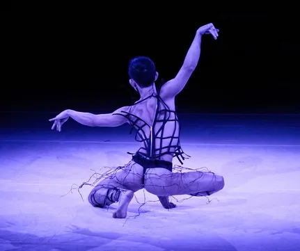 Minh sits in a toe squat, back to the camera, arms stretched in a dramatic pose, wearing a black G string and strappy top.